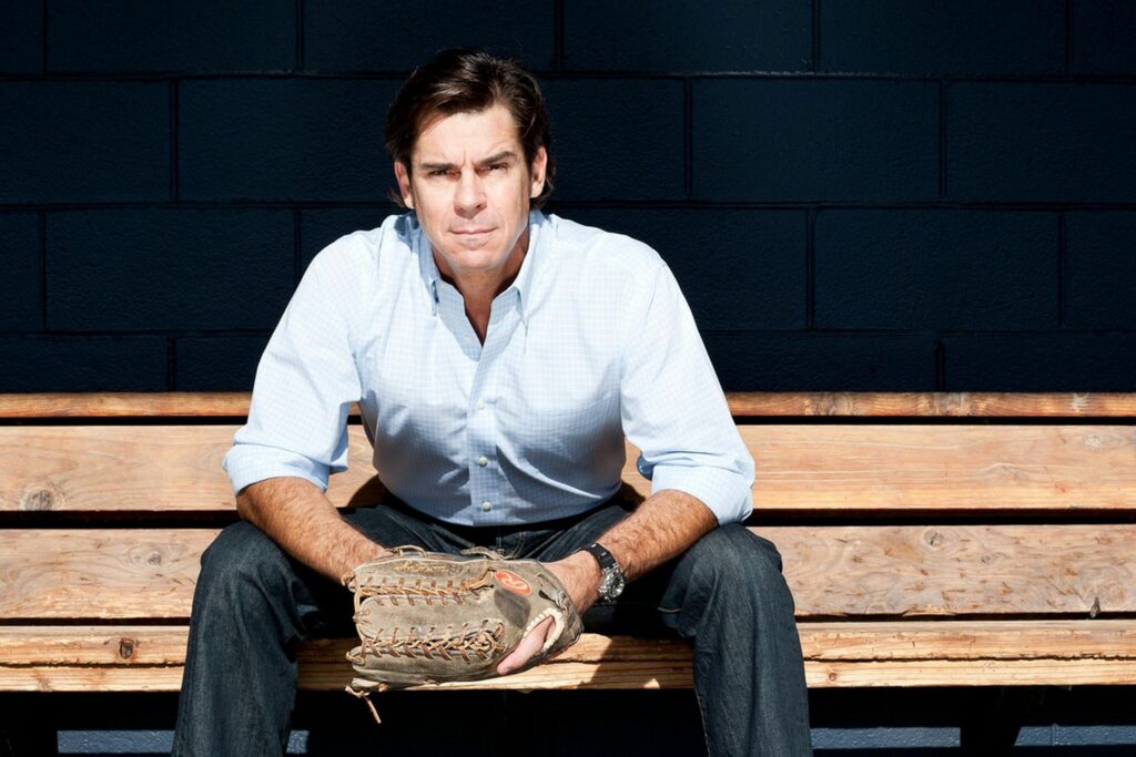 A man sitting on a wooden bench wearing a baseball glove on his left hand.